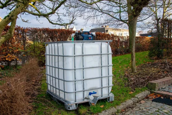 Gereinigde wit watertank van 1000 liter met gegalvaniseerde kooi, accu pomp, aftapkraan en onderstel in metaal of kunststof. linkerzijaanzicht-tuinomgeving-zonder-slang