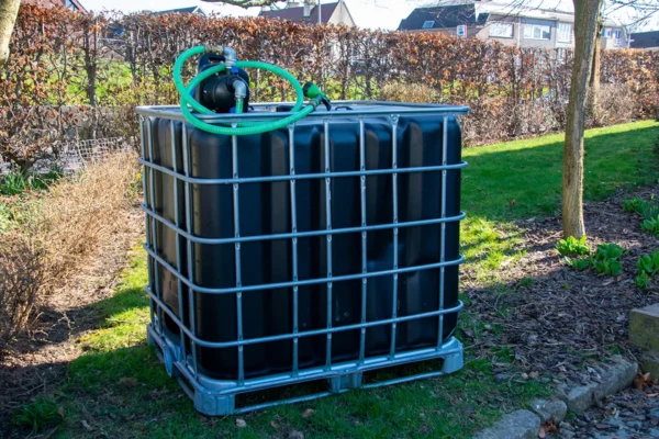 Vernieuwde zwarte watertank van 1000 liter met gegalvaniseerde kooi, pomp, aftapkraan en onderstel in metaal of kunststof. zijaanzicht-met-uitgaande-slang-in-tuinomgeving