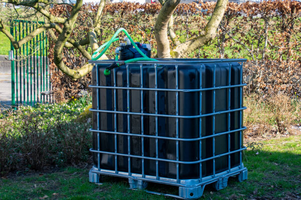 Vernieuwde zwarte watertank van 1000 liter met gegalvaniseerde kooi, accu pomp, aftapkraan en onderstel in metaal of kunststof. zijaanzicht-achteraanzicht-met-uitgaande-slang-in-tuinomgeving